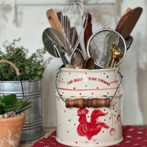 May include: A cream-coloured kitchen utensil holder with a red rooster design and the words "fait main tout naturel". The holder contains various kitchen tools, including whisks and wooden spoons. A small metal bucket and a terracotta pot are in the background.