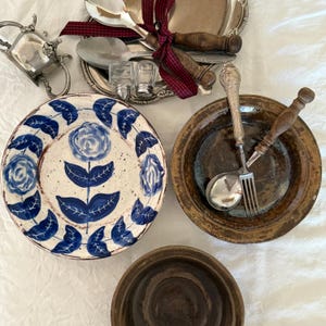 Op de afbeelding: Een verzameling vintage serviesgoed, waaronder een bord met blauwe bloemmotieven, zilveren serveeronderdelen en rustieke gebruiksvoorwerpen met houten handvatten. De opstelling omvat een zilveren serveerschaal, zout- en pepervaatjes en een kleine zilveren kan.
