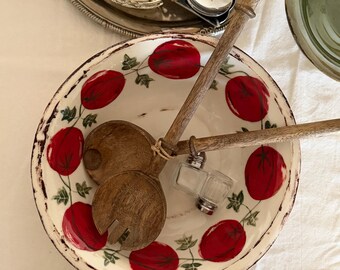 May include: A white ceramic bowl with a red tomato pattern and green leaves. Wooden salad servers and a glass salt and pepper shaker set rest inside the bowl. A silver serving tray with silverware is in the background.