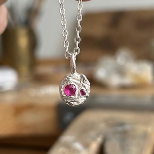 May include: A silver pendant necklace with a textured, orb-shaped charm. The charm has two bright red, round gemstones. The delicate silver chain complements the gemstones. The necklace is a piece of jewellery.