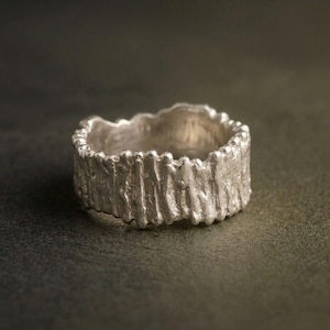Könnte beinhalten: Ein silberner Ring mit einer texturierten, unebenen Oberfläche, die an Baumrinde erinnert. Das Band weist vertikale Erhebungen und tiefe Rillen auf, die sich über den gesamten Umfang erstrecken und eine raue, organische Optik erzeugen.