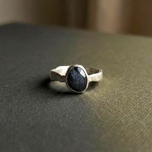 May include: A silver ring featuring a central oval-shaped, faceted dark blue gemstone. The metal band has a textured, uneven surface with a matte finish, holding the stone in a simple bezel setting that highlights the deep colour of the gem.