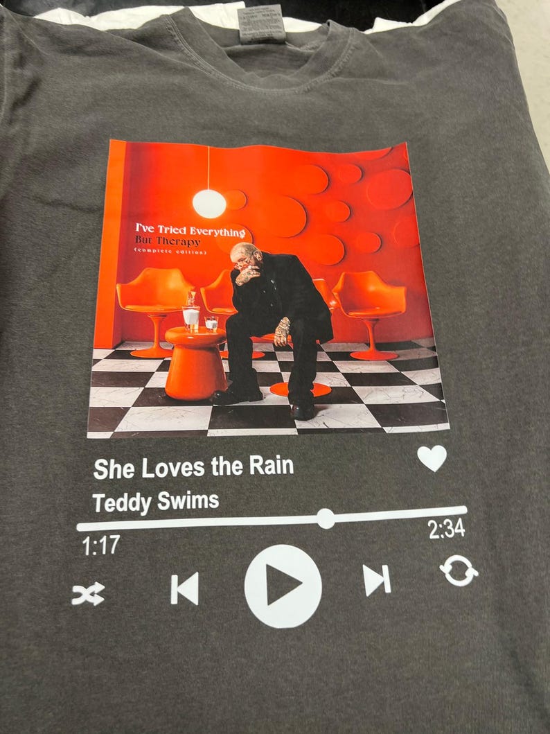 May include: Dark gray t-shirt featuring a graphic of a music player interface with the song title "She Loves the Rain" by Teddy Swims. The graphic includes an image of a person sitting in an orange room. The text "I've Tried Everything But Therapy" is also visible.