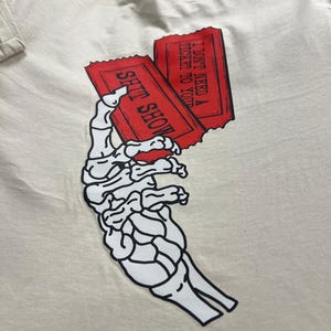 May include: Cream-colored t-shirt featuring a black and white skeleton hand holding two red tickets. The tickets read "SHIT SHOW" and "I DON'T NEED A TICKET TO YOUR". The shirt is made of a soft material.