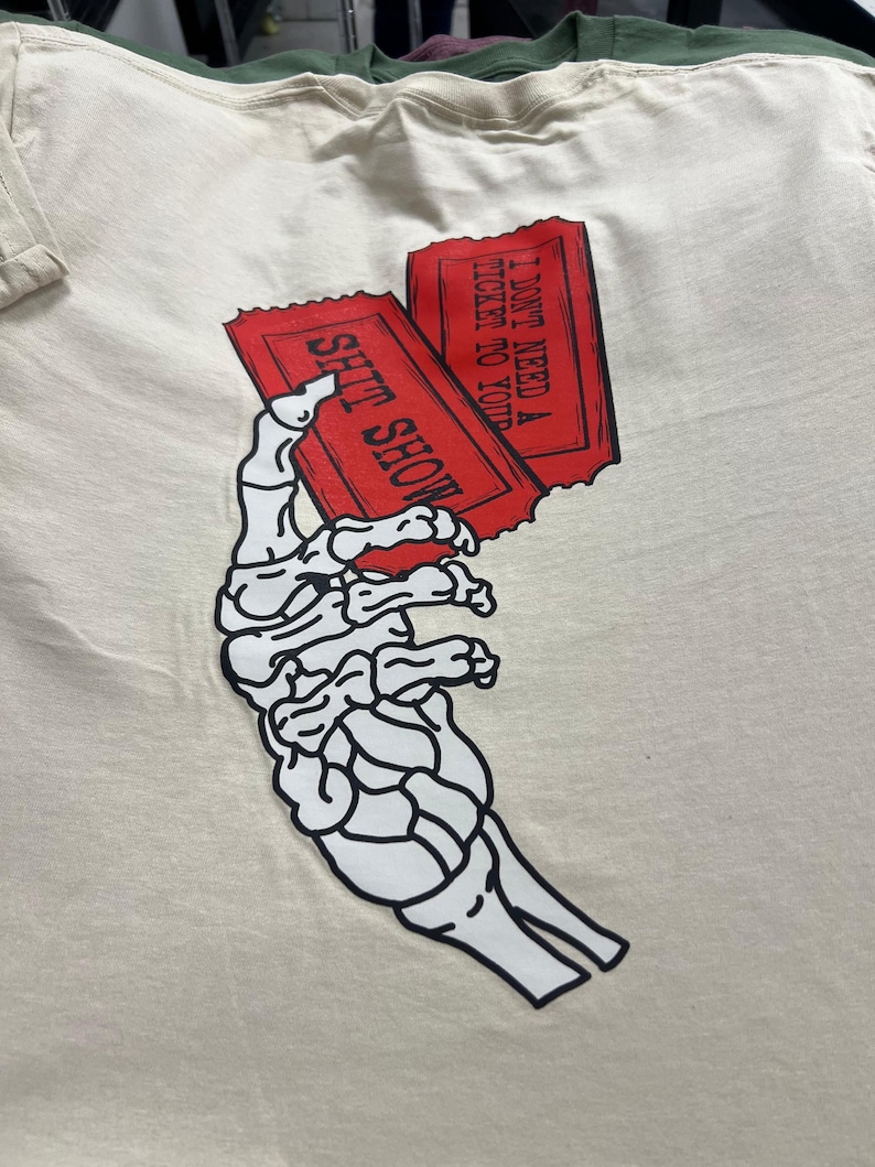 May include: Cream-colored t-shirt featuring a black and white skeleton hand holding two red tickets. The tickets read "SHIT SHOW" and "I DON'T NEED A TICKET TO YOUR". The shirt is made of a soft material.
