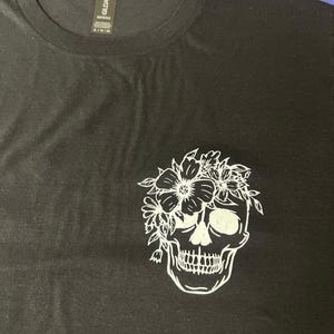 May include: Black t-shirt featuring a white floral skull graphic. The skull is detailed with eye sockets, teeth, and a floral crown. The shirt has a crew neck and short sleeves. The Gildan brand tag is visible.