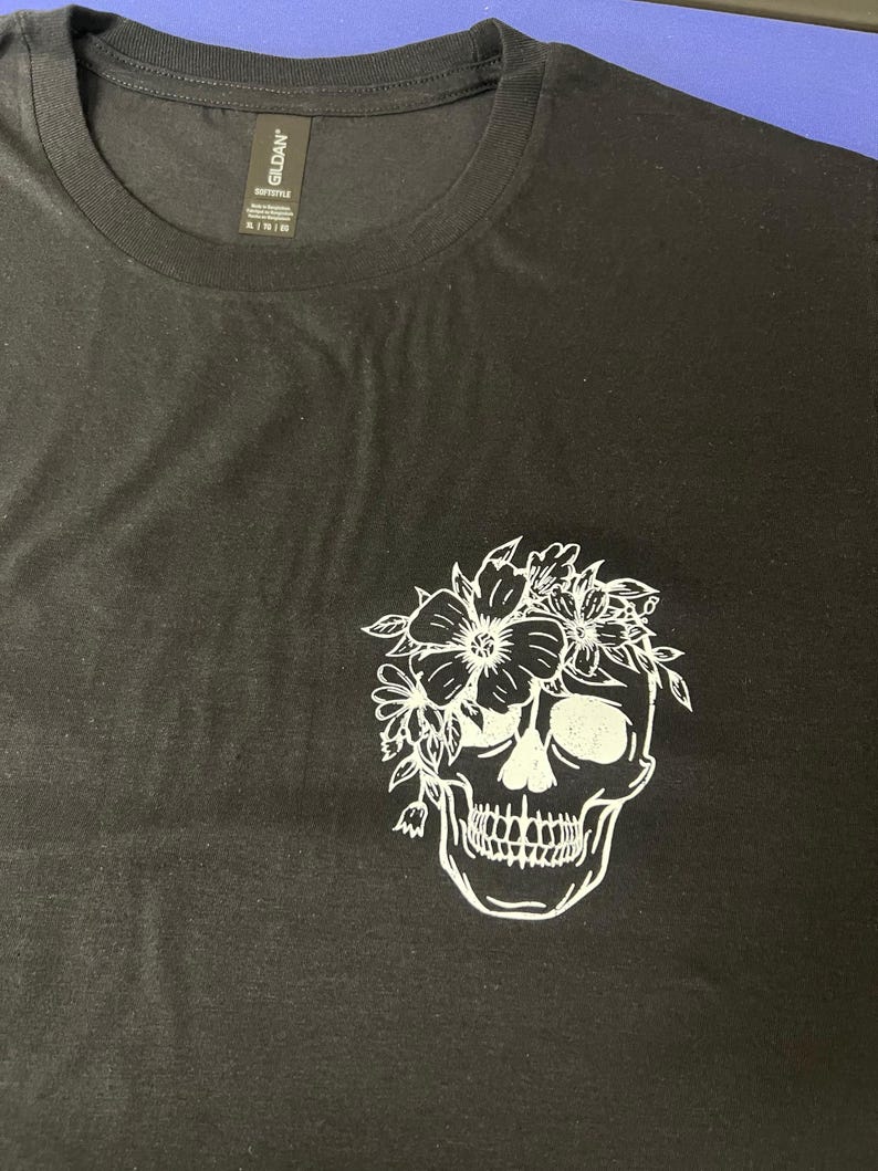 May include: Black t-shirt featuring a white floral skull graphic. The skull is detailed with eye sockets, teeth, and a floral crown. The shirt has a crew neck and short sleeves. The Gildan brand tag is visible.