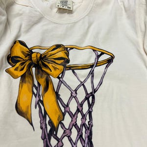 May include: Cream-colored t-shirt featuring a basketball net design with a large yellow bow. The net is purple, and the bow has yellow and black shading. The shirt has a crew neck and short sleeves. The tag reads "Comfort Colors".