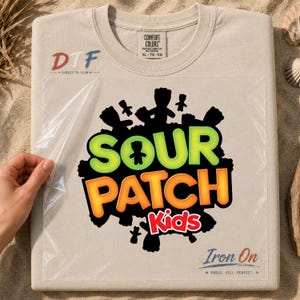 May include: A beige t-shirt featuring a Sour Patch Kids graphic transfer with a clear plastic film overlay. The design includes the text SOUR PATCH Kids in bright green, orange, and red, surrounded by black candy silhouettes and DTF branding.