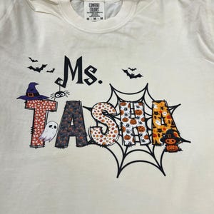 May include: Cream-colored t-shirt with the text "Ms. TASHA" in a Halloween-themed design. The letters are decorated with patterns of bats, pumpkins, and a witch's hat. A spider and ghost are also included.