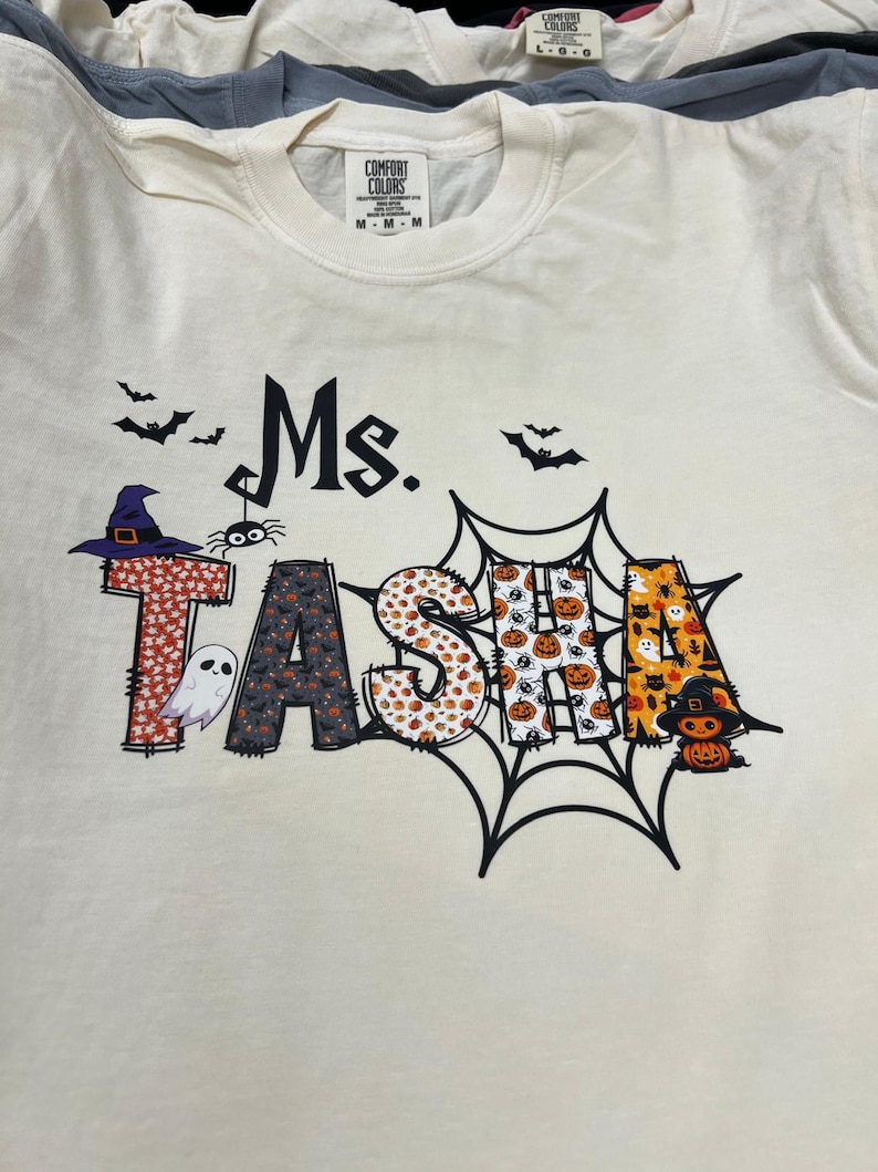 May include: Cream-colored t-shirt with the text "Ms. TASHA" in a Halloween-themed design. The letters are decorated with patterns of bats, pumpkins, and a witch's hat. A spider and ghost are also included.