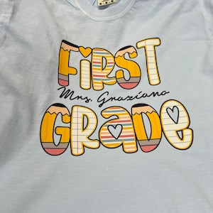 May include: Light blue t-shirt with the words "FIRST GRADE Mrs. Graziano" in a colorful pencil-themed design. The text is in a playful font with hearts and lined paper details, perfect for back-to-school.