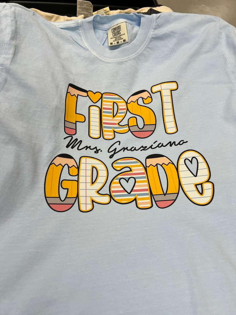 May include: Light blue t-shirt with the words "FIRST GRADE Mrs. Graziano" in a colorful pencil-themed design. The text is in a playful font with hearts and lined paper details, perfect for back-to-school.