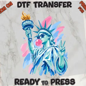May include: A DTF transfer print featuring the Statue of Liberty wearing sunglasses and blowing a pink bubble. The statue holds a torch with yellow flames and makes a peace sign. Text reads IRON ON DTF TRANSFER PRINT FOR READY TO PRESS.