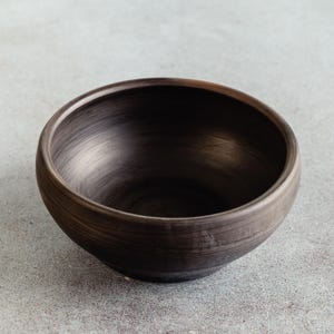 Peut inclure: Un bol en céramique brun foncé de forme arrondie. Le bol a une finition lisse et polie avec une texture visible et une large ouverture. L'intérieur est d'une couleur sombre et profonde, contrastant avec le bord plus clair.