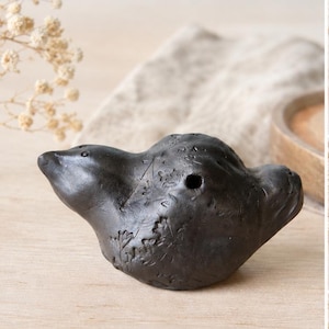 Pode incluir: Ocarina de cerâmica castanha escura em forma de pássaro. O instrumento tem um acabamento liso e fosco com entalhes em forma de folha e um único orifício. Repousa sobre uma superfície de madeira clara.