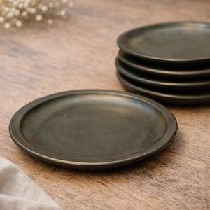 Op de afbeelding: Een set van zes donkergrijze keramische borden met een licht verhoogde rand. De borden zijn rond en hebben een matte afwerking. De borden staan op een houten oppervlak.