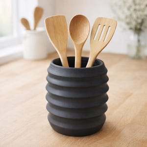 May include: A black, ribbed ceramic utensil holder with three wooden kitchen tools: a spatula, a spoon, and a slotted spatula. The utensils are light brown, and the holder is cylindrical. Additional utensils are visible in the background.