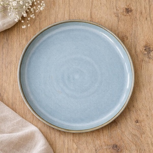 Op de afbeelding: Een ronde, lichtblauwe keramische plaat met een licht getextureerd oppervlak en een dunne, bruine rand. De plaat staat op een houten oppervlak, met een beige doek en witte bloemen op de achtergrond.