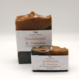 May include: Two bars of handmade soap with a brown and black color scheme. The soap is labeled "Sandalwood & Vanilla" and made with rice oil and shea butter. The smaller bar is 1.7 oz / 48 g.