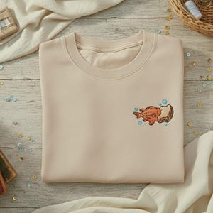 May include: A beige crewneck sweatshirt with an embroidered design of a cartoon fish and bread. The sweatshirt is folded on a light wooden surface, with fabric, books, and sewing supplies in the background.