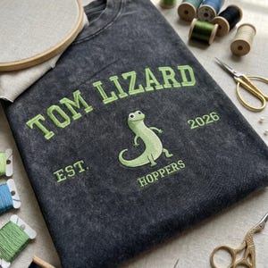 Puede incluir: Camiseta gris oscuro con el texto "TOM LIZARD" en verde, un gráfico de lagarto y el texto "EST. HOPPERS 2026." Suministros de bordado, incluyendo carretes de hilo, tijeras y un bastidor de bordado, están dispuestos alrededor de la camiseta.