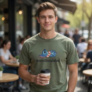 May include: Olive green t-shirt featuring a blue cartoon character with a floral lei, with the word "overstimulated" in black text. The person is holding a coffee cup with a brown sleeve.