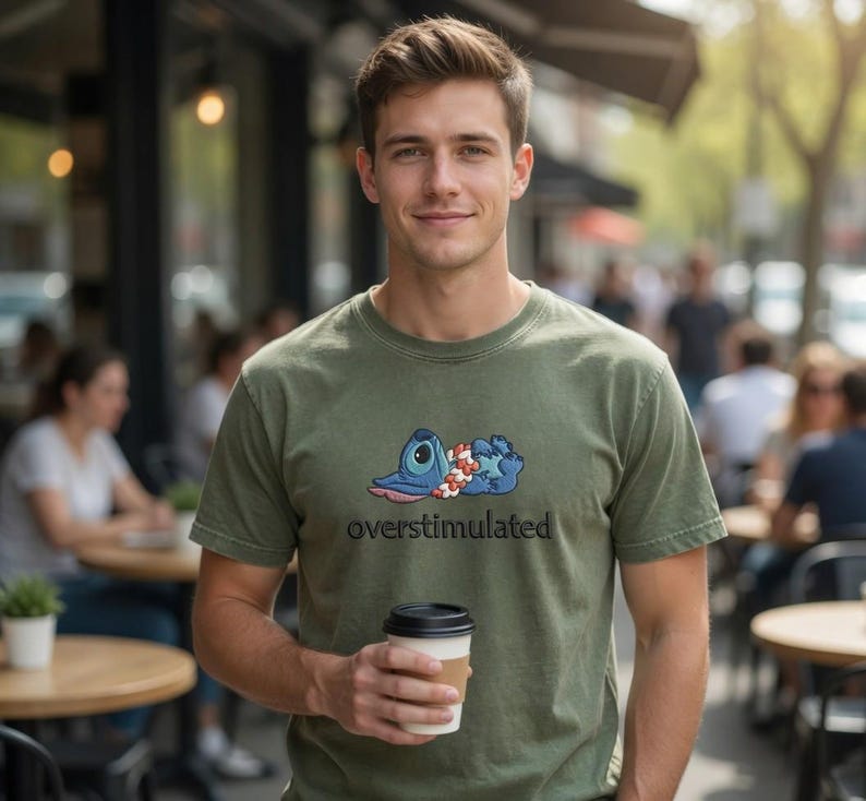 May include: Olive green t-shirt featuring a blue cartoon character with a floral lei, with the word "overstimulated" in black text. The person is holding a coffee cup with a brown sleeve.