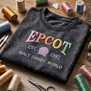 May include: A charcoal grey t-shirt featuring the embroidered word "EPCOT" in pastel shades. Below the word are the words "EST. 1982" and "WALT DISNEY WORLD". The shirt is surrounded by spools of thread, scissors, and sewing notions on a wooden surface.
