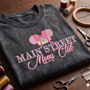 Puede incluir: Camiseta gris oscuro con el texto bordado "MAIN STREET Moms Club". El diseño incluye un lazo rosa y orejas de ratón. Suministros de costura, incluyendo carretes de hilo, tijeras y un bastidor, están dispuestos alrededor de la camiseta.
