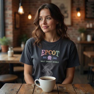 May include: A dark grey t-shirt featuring the word "EPCOT" in colourful embroidery, with "EST. 1982" and "WALT DISNEY WORLD" below. A white mug of coffee sits on a wooden table. The image is taken in a cafe setting.