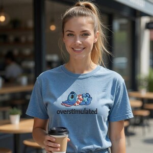 May include: A light blue t-shirt featuring a cartoon character with the word "overstimulated" in black text. The cartoon character is blue with large eyes and is lying down. The person in the image is holding a coffee cup.
