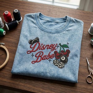Puede incluir: Camiseta azul claro con la frase bordada "Disney Babe" en rojo. El dise&ntilde;o incluye cerezas, dados y una silueta de Mickey Mouse. Hilos de bordar y tijeras son visibles en el fondo.