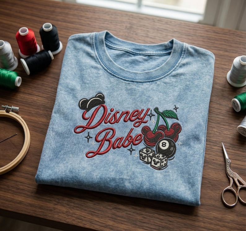Puede incluir: Camiseta azul claro con la frase bordada "Disney Babe" en rojo. El dise&ntilde;o incluye cerezas, dados y una silueta de Mickey Mouse. Hilos de bordar y tijeras son visibles en el fondo.
