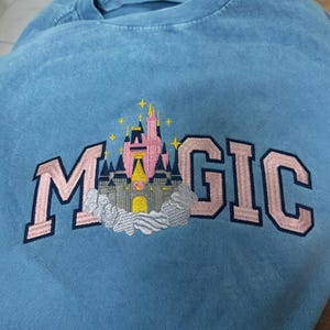May include: A light blue sweatshirt with the word "MAGIC" embroidered in pink block letters. A pink and gray castle design is embroidered in the center of the word "MAGIC". The sweatshirt is made of a soft material.