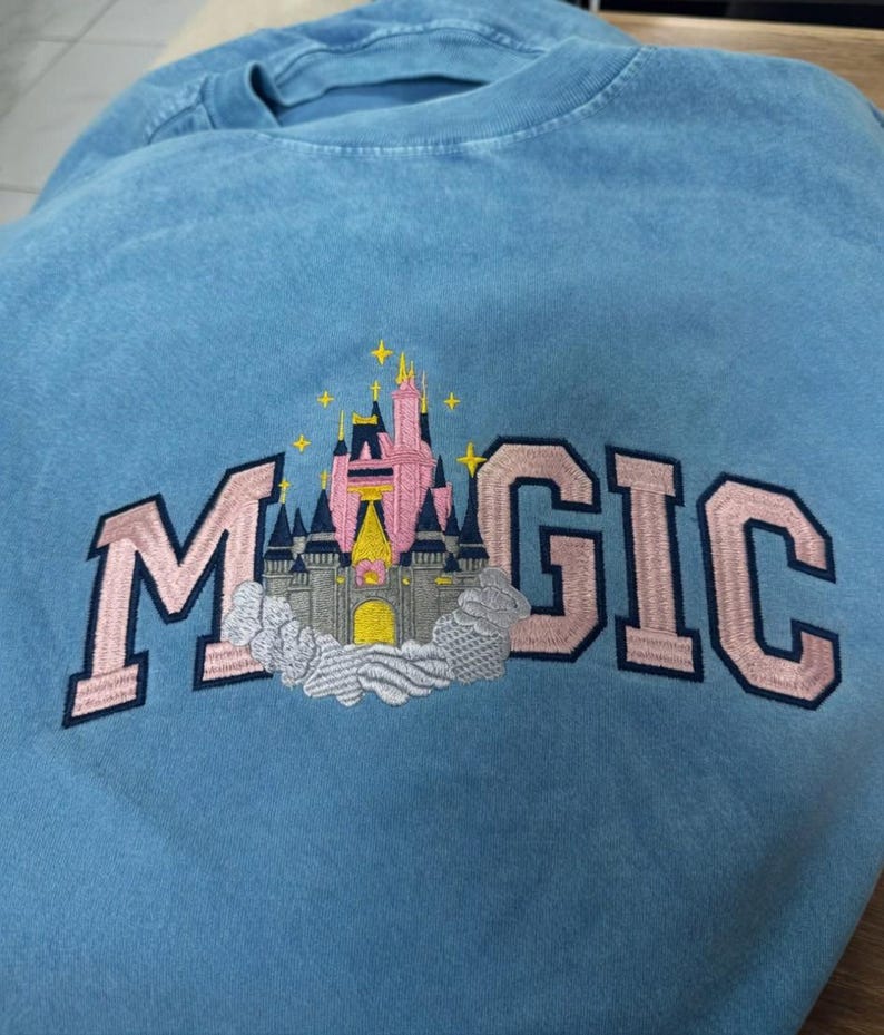 May include: A light blue sweatshirt with the word "MAGIC" embroidered in pink block letters. A pink and gray castle design is embroidered in the center of the word "MAGIC". The sweatshirt is made of a soft material.