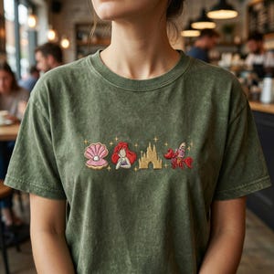 K&ouml;nnte beinhalten: Olivgr&uuml;nes T-Shirt mit gestickten Cartoon-Bildern einer Muschel, einer Meerjungfrau, eines Schlosses und einer Krabbe. Das Shirt hat einen Rundhalsausschnitt und kurze &Auml;rmel. Die Stickerei ist in den Farbt&ouml;nen Rosa, Rot, Gold und Orange gehalten.