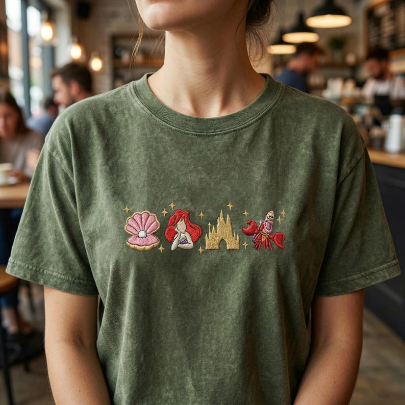 K&ouml;nnte beinhalten: Olivgr&uuml;nes T-Shirt mit gestickten Cartoon-Bildern einer Muschel, einer Meerjungfrau, eines Schlosses und einer Krabbe. Das Shirt hat einen Rundhalsausschnitt und kurze &Auml;rmel. Die Stickerei ist in den Farbt&ouml;nen Rosa, Rot, Gold und Orange gehalten.
