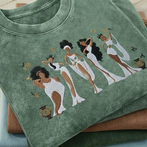 Peut inclure: T-shirt vert avec un motif brod&eacute; de quatre personnages stylis&eacute;s en robes blanches. Les personnages ont les cheveux fonc&eacute;s et sont dans diverses poses. Le t-shirt est pli&eacute; sur d'autres chemises pli&eacute;es dans des tons de marron et de bleu.