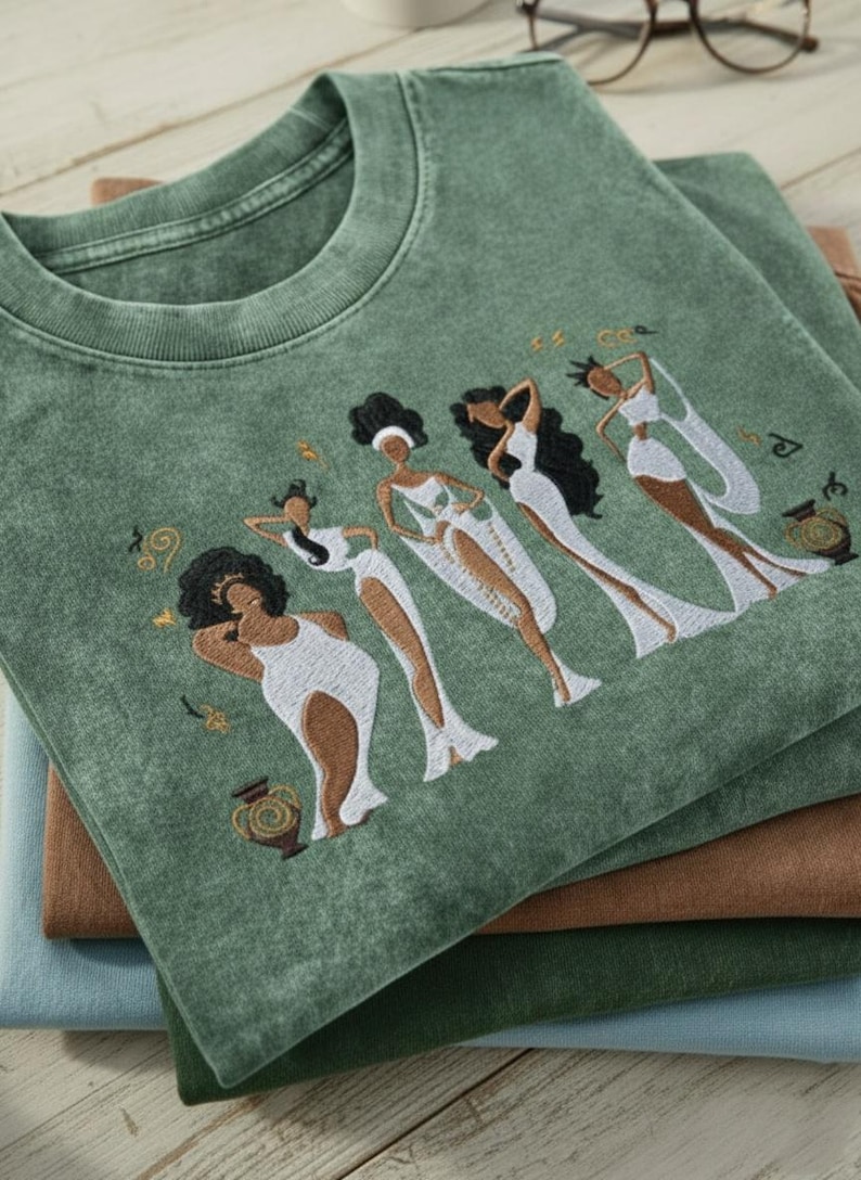 Peut inclure: T-shirt vert avec un motif brod&eacute; de quatre personnages stylis&eacute;s en robes blanches. Les personnages ont les cheveux fonc&eacute;s et sont dans diverses poses. Le t-shirt est pli&eacute; sur d'autres chemises pli&eacute;es dans des tons de marron et de bleu.