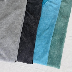 K&ouml;nnte beinhalten: Nahaufnahme von vier gefalteten T-Shirts in verschiedenen Farben: Grau, Schwarz, T&uuml;rkis und Gr&uuml;n. Die Shirts haben ein strukturiertes, verwaschenes Aussehen, was auf einen Vintage- oder gewaschenen Stil hindeutet. Der Stoff wirkt weich und bequem.