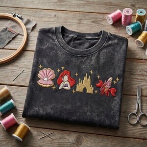 K&ouml;nnte beinhalten: Dunkelgraues T-Shirt mit gesticktem Design einer Muschel, einer Meerjungfrau, einem Schloss und einer Krabbe. Die Stickerei ist in Rosa-, Rot-, Gold- und Wei&szlig;t&ouml;nen gehalten. N&auml;hutensilien sind im Bild zu sehen.