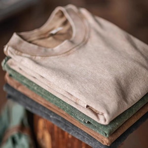 May include: A stack of folded t-shirts in various neutral colors: beige, green, brown, and gray. The shirts have a vintage, worn look, with a classic crew neck design. The fabric appears soft and slightly textured, suggesting a comfortable, casual style.
