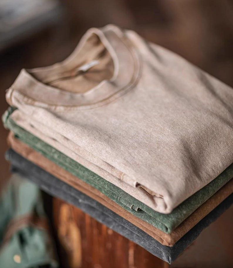 May include: A stack of folded t-shirts in various neutral colors: beige, green, brown, and gray. The shirts have a vintage, worn look, with a classic crew neck design. The fabric appears soft and slightly textured, suggesting a comfortable, casual style.