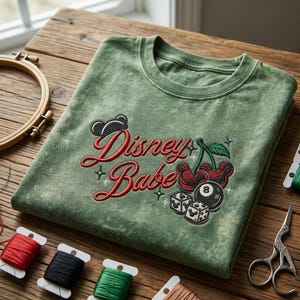 Puede incluir: Una camiseta verde salvia con la inscripci&oacute;n bordada "Disney Babe" en escritura roja. El dise&ntilde;o incluye cerezas, dados y una silueta de Mickey Mouse. La camiseta est&aacute; doblada sobre una superficie de madera.