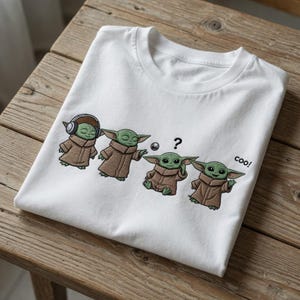 Puede incluir: Camiseta blanca con un dise&ntilde;o de dibujos animados de un personaje verde con aspecto de extraterrestre con una t&uacute;nica marr&oacute;n. El personaje se muestra en cuatro poses diferentes, con la palabra "coo!" impresa.