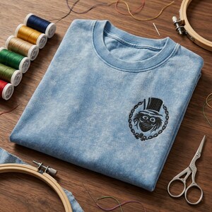 Op de afbeelding: Lichtblauw T-shirt met ronde hals en een zwart geborduurd ontwerp van een schedel met hoge hoed. Naaibenodigdheden, waaronder garenklosjes, scharen en borduurringen, omringen het T-shirt.