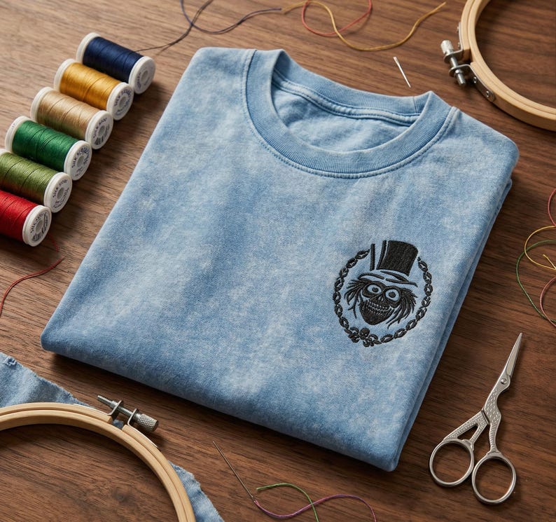 Op de afbeelding: Lichtblauw T-shirt met ronde hals en een zwart geborduurd ontwerp van een schedel met hoge hoed. Naaibenodigdheden, waaronder garenklosjes, scharen en borduurringen, omringen het T-shirt.