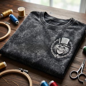 Op de afbeelding: Donkergrijs t-shirt met een wit geborduurd ontwerp van een schedel met hoge hoed, omringd door een decoratieve krans. Het shirt is opgevouwen op een houten oppervlak, met naaibenodigdheden zoals garenklosjes, borduurringen en een schaar.
