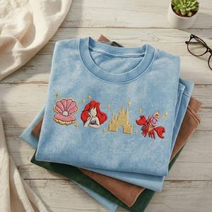 K&ouml;nnte beinhalten: Hellblaues T-Shirt mit gestickten Bildern einer Muschel, einer rothaarigen Frau, einem Schloss und einer roten Krabbe. Das Shirt ist auf anderen gefalteten Shirts in Gr&uuml;n- und Braunt&ouml;nen gefaltet.